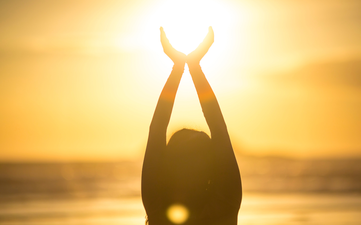 Silhouette of a person raising their hands as if to hold the sun as it rises over the ocean